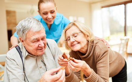 Ein älterer Mann und eine ältere Frau beschäftigen sich miteinander. Im Hintergrund steht die Pflegerin und lacht.